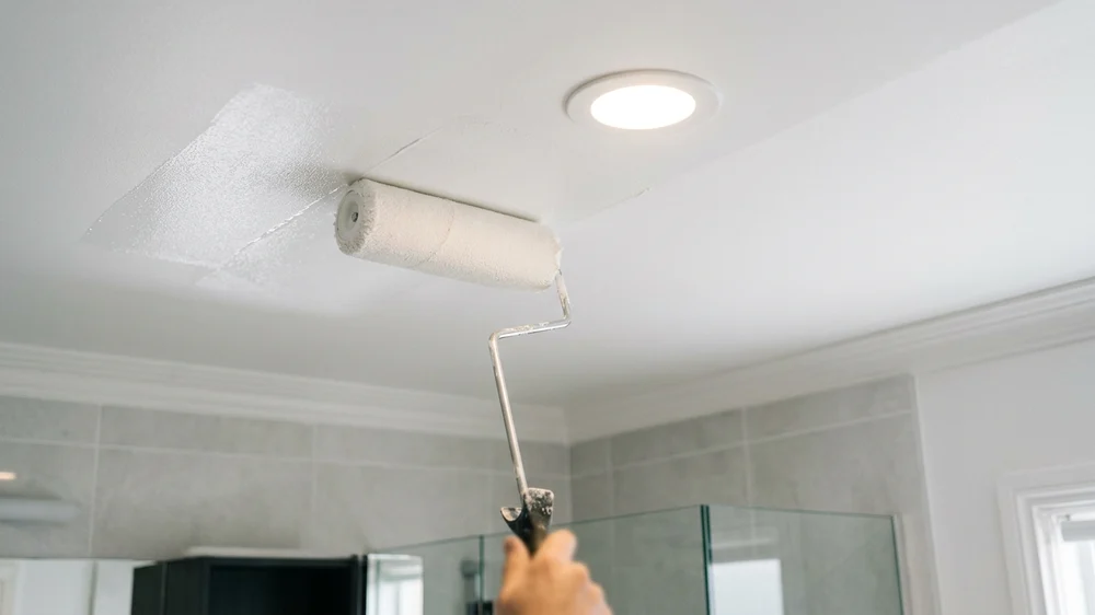 Applying smooth matte white mold-resistant paint to a bathroom ceiling with a paint roller